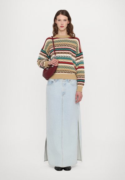 Maglione lavorato a maglia con motivi multicolori, abbinato a una gonna maxi in denim blu chiaro con spacchi laterali. La modella tiene una piccola borsa bordeaux.