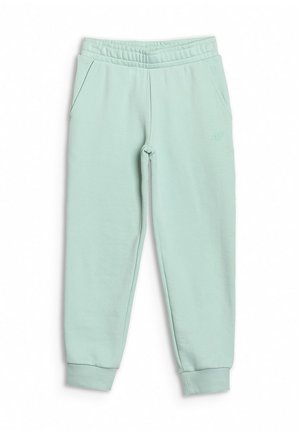 Młoda osoba z długimi blond włosami, ubraną w fioletowy t-shirt, miętowe spodnie dresowe, białe skarpetki oraz jasnozielone i białe buty sportowe, uśmiechającą się.