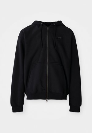Črna zip-up kapuca izdelana iz mehkega materiala, ki ima kapuco z vrvico, dolge rokave, sprednji kenguru žep in majhen bel logo na prsih.