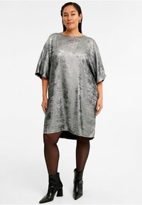 Silberne, metallische Kleid mit lockerer Passform, kurzen Ärmeln und asymmetrischem Saum. Kombiniert mit schwarzen Knöchelstiefeln und transparenten schwarzen Strumpfhosen.