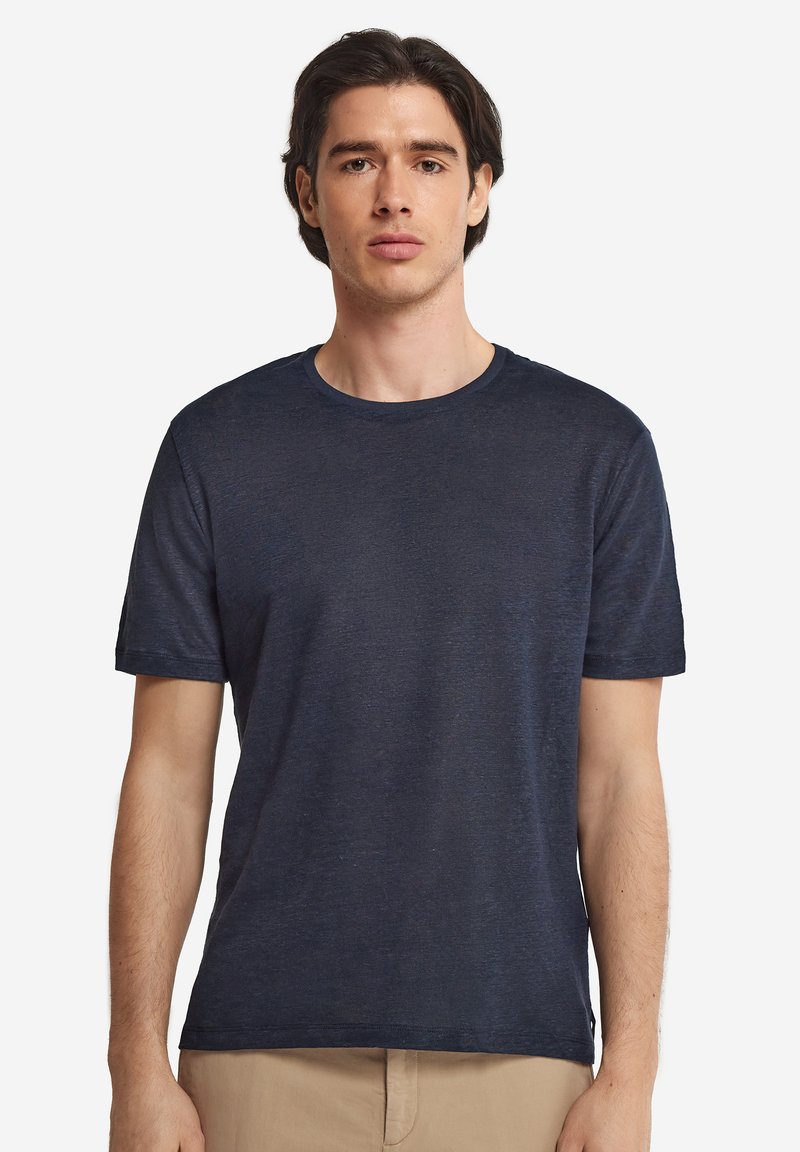 Jeune homme aux cheveux mi-longs foncés portant un t-shirt bleu foncé uni à manches courtes et un pantalon beige, faisant face à l'avant.