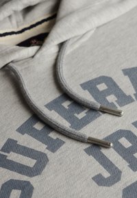 Close-up van een grijze hoodie met donkerblauwe blokletters en bijpassende grijze koorden met metalen uiteinden.