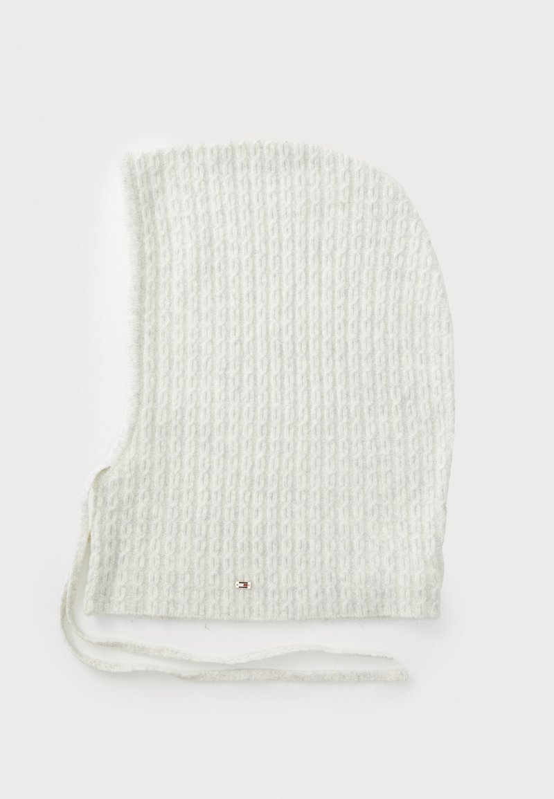 Fehér kötött sapka texturált mintával. Nyaknál megkötőkkel és egy kis fémszerelvénnyel ellátva. Puha, rugalmas anyagú, íves formájú.