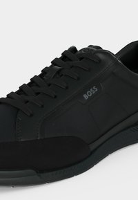 Baskets basses noires avec tissu texturé et panneaux en cuir, lacets noirs et logo "BOSS" sur le côté.
