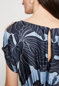 Blouse à motif floral bleu marine avec une finition satinée, dotée d'un col rond, de manches courtes et d'un détail en forme de trou de serrure à l'arrière.