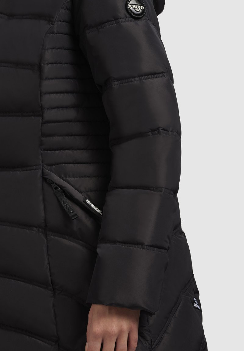 Manche de veste d'hiver noire rembourrée avec un patch logo "Khujo" sur le haut du bras et une poche zippée près du poignet sur un fond clair.