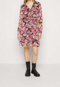 Blommig klänning med rosa och vita blommor på en mörk bakgrund, långa ärmar, V-ringning och midjebälte, i kombination med svarta ankellånga stövlar.