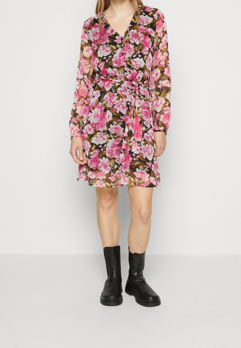 Blommig klänning med rosa och vita blommor på en mörk bakgrund, långa ärmar, V-ringning och midjebälte, i kombination med svarta ankellånga stövlar.