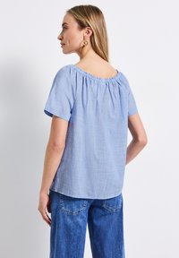 Blusa azzurro chiaro a maniche corte con scollatura arricciata e una sottile trama, abbinata a jeans in denim blu. Nessun motivo o accentuazione visibile.