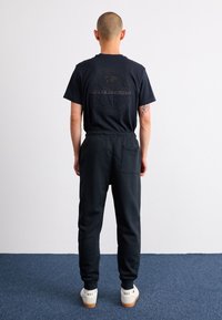 Svart t-shirt med kort ärm och rund halsringning, med präglad logotyp på baksidan, ihop med svarta avsmalnande joggers och vita sneakers.