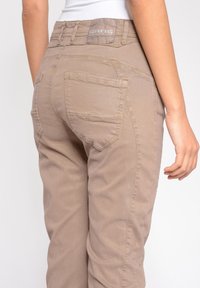 Beige bavlněn�é kalhoty s mírně zvýšeným pasem, výraznými zadními kapsami a označením na pásku. Hladká textura s přizpůsobeným střihem.