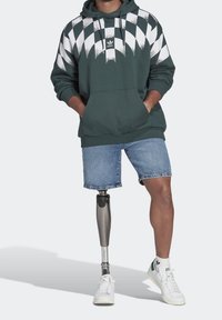 Person som bär en grön och vit mönstrad hoodie, denimshorts, vita sneakers och en protes på vänsterbenet, stående med händerna i fickorna.