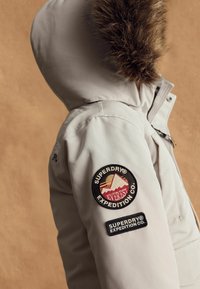 Parka gri deschis cu o glugă căptușită cu blană sintetică. Prezentă logo-ul brodat cu munte și semnele brand-ului pe mânecă, textură netedă.