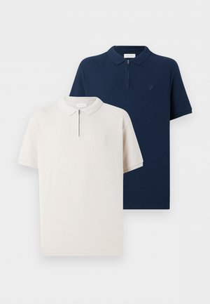 Férfi áll, bézs rövid ujjú galléros pólót, kék farmert, fekete cipőt és karórát visel, sima fehér háttér előtt.