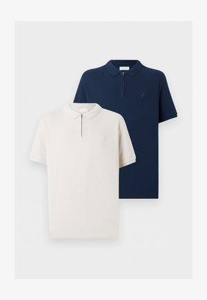 Άνδρας όρθιος που φοράει μπεζ πόλο με κοντά μανίκια, μπλε τζιν, μαύρα παπούτσια και ρολόι χειρός μπροστά σε απλό λευκό φόντο.