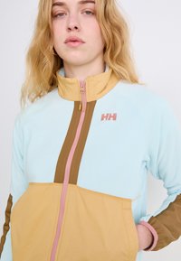 Jachetă din fleece cu un corp de culoare albastru deschis, panou frontal maro, fermoar roz și guler înalt. Logo pe piept; mâneci lungi cu accente maronii.