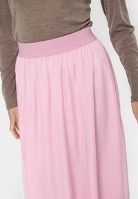 Gonna plissettata rosa chiaro con fascia elastica, realizzata in tessuto morbido e fluido, mostrata parzialmente accanto a una maglia a maniche lunghe marrone.