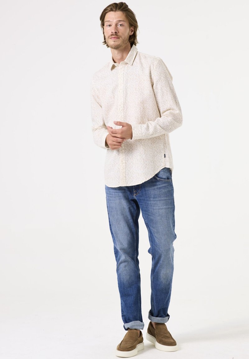 Camisa de botón de manga larga color crema con un patrón floral, combinada con jeans azules y zapatos marrones sin cordones. Estilo casual, ajuste relajado.