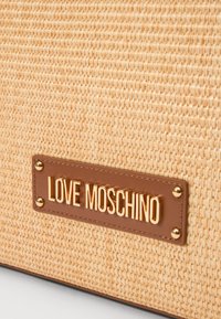 Borsa intrecciata color beige con un'etichetta in pelle marrone, decorata con la scritta "LOVE MOSCHINO" in oro, arricchita da borchie dorate. Dettagli della superficie texture visibili.