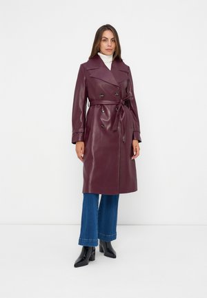 Femme debout portant un long trench-coat en cuir bordeaux noué à la taille, un jean bleu, des bottes noires et un pull à col roulé blanc.
