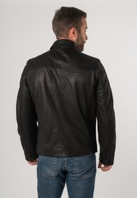 Giubbotto in pelle nera con finitura testurizzata, collo alto e vestibilità su misura. Sono visibili le cerniere sui polsini e dettagli di cucitura discreti.
