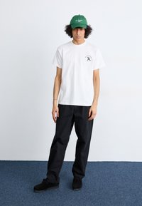 White cotton t-shirt with black graphic, black pants, and green cap with white text. Model stands on a blue carpet against a neutral background.