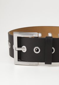 Czarny skórzany pasek z srebrną klamrą, ozdobiony okrągłymi otworami. Wewnętrzna strona jest w kolorze jasnobrązowym. Logo jest wytłoczone na klamrze.