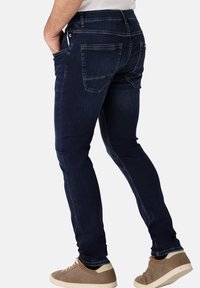 Dunkelblaue Denim-Jeans mit taillierter Passform, fünf Taschen und dezenten Stickdetails, kombiniert mit hellbraunen Sneakers.