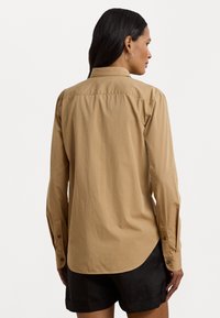Camicia button-up marrone chiaro, maniche lunghe, dettaglio sul retro, orlo curvo e texture liscia. Indossata con pantaloni corti neri.