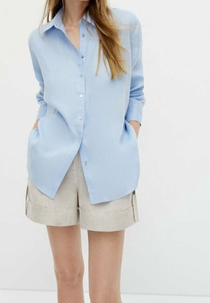 Personne portant une chemise bleu clair boutonnée à manches longues et un short beige avec poches, debout devant un fond blanc uni.