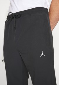Zwarte sportbroek met een elastische tailleband, voorzien van een trekkoord, zijzakken en een wit Jumpman-logo op de linker bovenbeen.