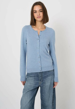 Frau trägt eine hellblaue, geknöpfte Strickjacke und weit geschnittene blaue Jeans, steht vor einem schlichten hellen Hintergrund.