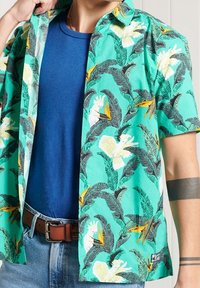 Camicia a maniche corte turchese con motivo di uccelli tropicali e foglie. Realizzata in tessuto leggero, presenta una chiusura frontale con bottoni e una texture liscia.