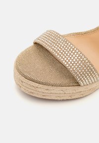 Sandalia de plataforma en color dorado con una amplia correa texturizada adornada con pequeños embellishments brillantes y una suela envuelta en yute.