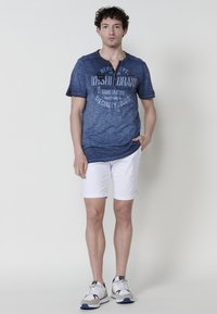 T-paita tummansinisestä kankaasta, lyhyet hihat, teksturoitu gradientti ja painettu logo, yhdistetty valkoisiin käärittyihin shortsit ja harmaisiin lenkkareihin. Yksinkertainen muotoilu.