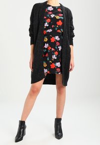 Cárdigan largo de punto negro sobre un vestido de estampado floral con flores rojas, naranjas y moradas sobre fondo negro. Botines negros hasta el tobillo.
