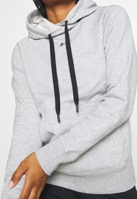 Sudadera gris de tejido suave con un bolsillo canguro, cordones negros y puños y dobladillo acanalados; presenta un pequeño logo en el pecho.