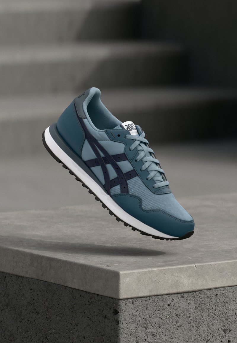 Kék és sötét tengerészkék sportcipő, fehér talppal, amely egy betonperem felett lebeg, a háttérben szürke lépcső blurjével.