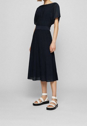 Femme portant une robe midi bleu marine à manches courtes et taille cintrée, associée à des sandales blanches plates avec des lanières aux chevilles.