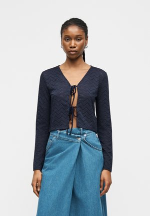 Kvinde med flettet hår iført navy langærmet crop top med knuder foran og højtaljede, vidde blå jeans mod hvid baggrund.