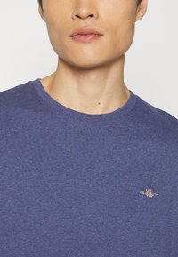 Nahaufnahme einer Person, die ein schlichtes blaues Rundhals-Shirt mit einem kleinen bestickten Emblem auf der linken Brust trägt.