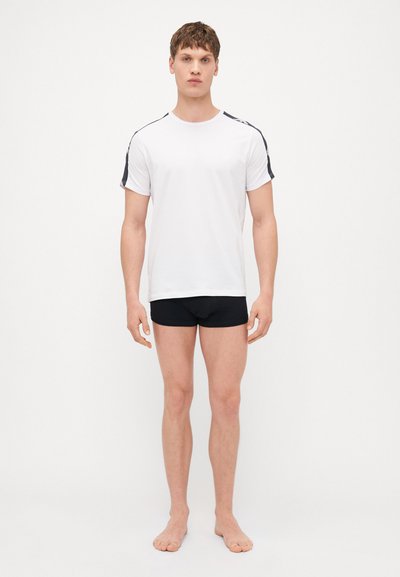 White short-sleeve t-shirt with black accents on the shoulders, paired with black fitted shorts. Model stands barefoot against a plain background.