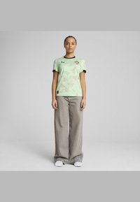 Jersey vert clair à manches courtes avec un motif subtil, accents noirs et logo brodé ; associé à un large pantalon gris et des chaussures blanches.