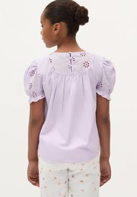 Helle lavendelfarbene Bluse mit bestickten Puffärmeln, detailliertem Ausschnitt und Knopfleiste im Rücken. Glattes Material, lockere Passform.