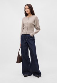 Μπεζ ζακέτα από μαλλί με κουμπιά μπροστά, συνδυασμένη με φαρδιά σκούρα τζιν παντελόνια. Το μοντέλο κρατά μια καφέ τσάντα, στέκεται μπροστά σε ανοιχτό γκρίζο φόντο.