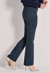 Vrouw in gestreepte marineblauwe broek en beige hoge hakken die tegen een effen lichte muur loopt.