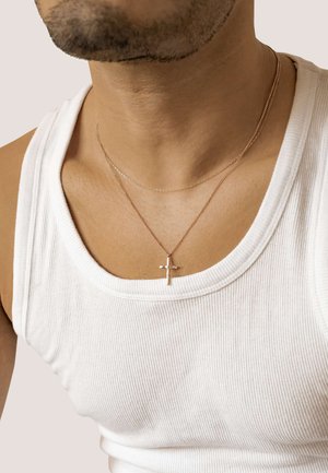 Goldfarbene Kreuzanhänger-Halskette, die über einem weißen Rippen-Top getragen wird, mit einer zartem silbernen Kette, die darunter geschichtet ist.