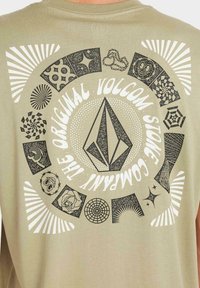 Camiseta de algodón beige que presenta un diseño gráfico circular con ilustraciones en blanco y negro, incluyendo formas y patrones geométricos, y un logo centrado.