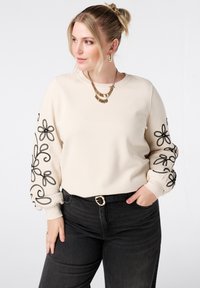 Sweat-shirt beige avec broderies florales noires sur les manches. Doté d'un col rond et de poignets côtelés. Associé à un jean foncé et une ceinture noire.