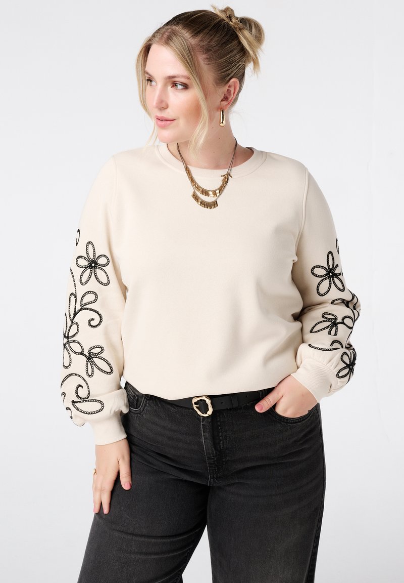 Sweat-shirt beige avec broderies florales noires sur les manches. Doté d'un col rond et de poignets côtelés. Associé à un jean foncé et une ceinture noire.